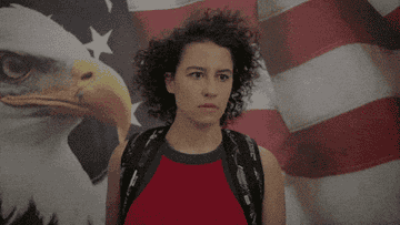 Ilana Glazer standing in front of an American Flag giving a salute