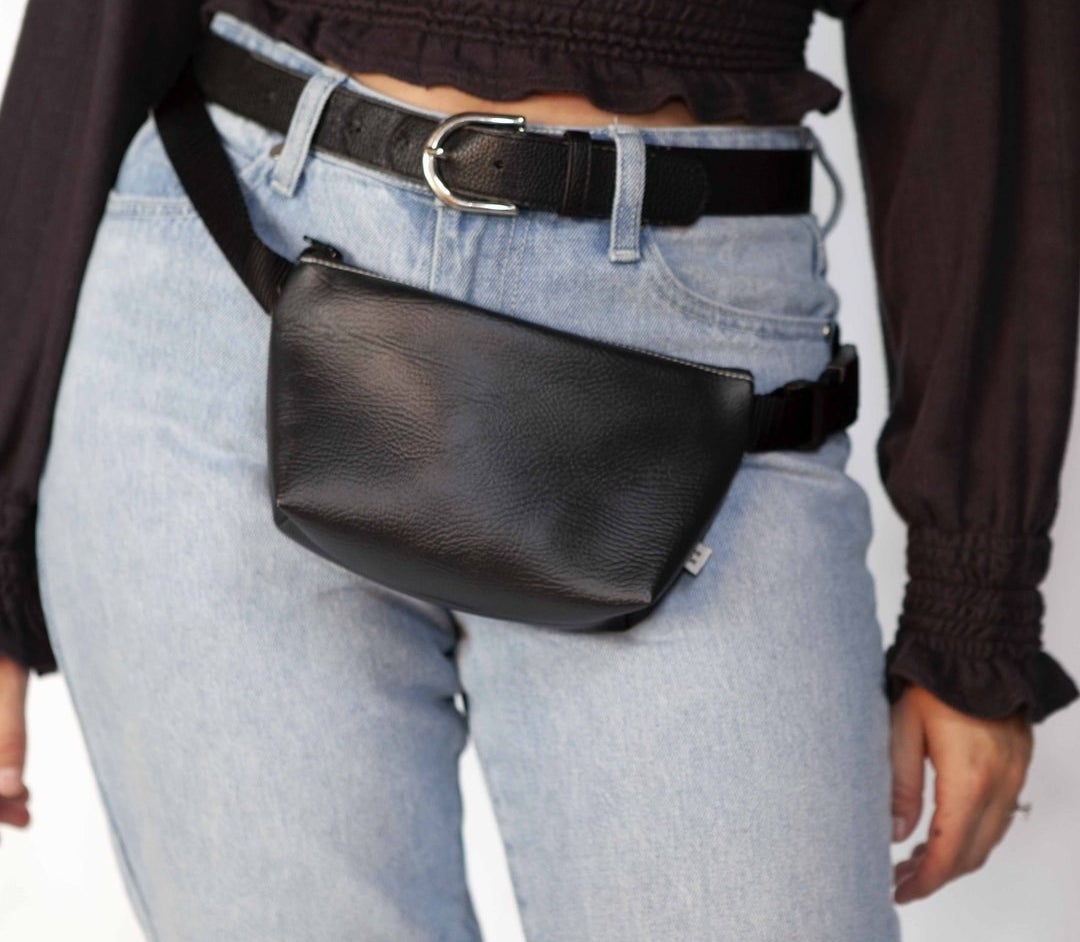 Model is wearing denim jeans, a black top, and a black belt bag around their hips