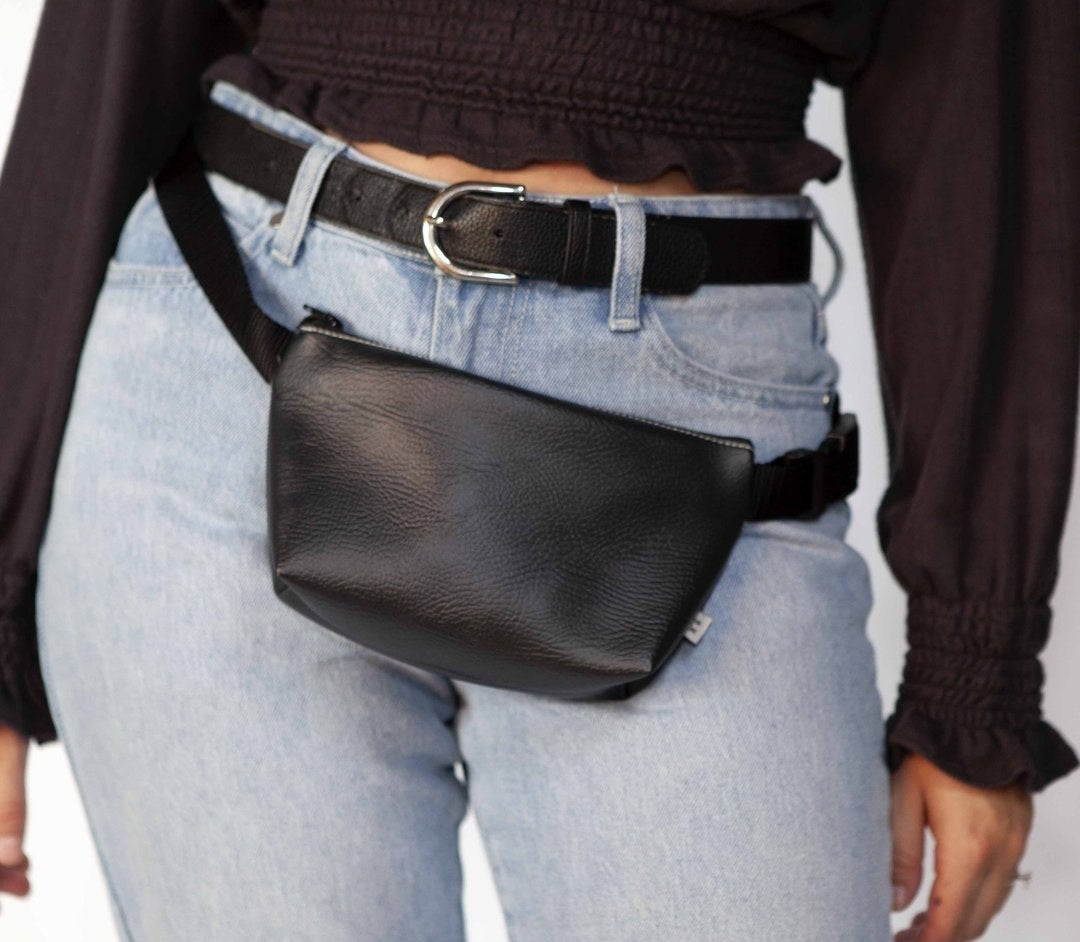 Model is wearing denim jeans, a black top, and a black belt bag around their hips
