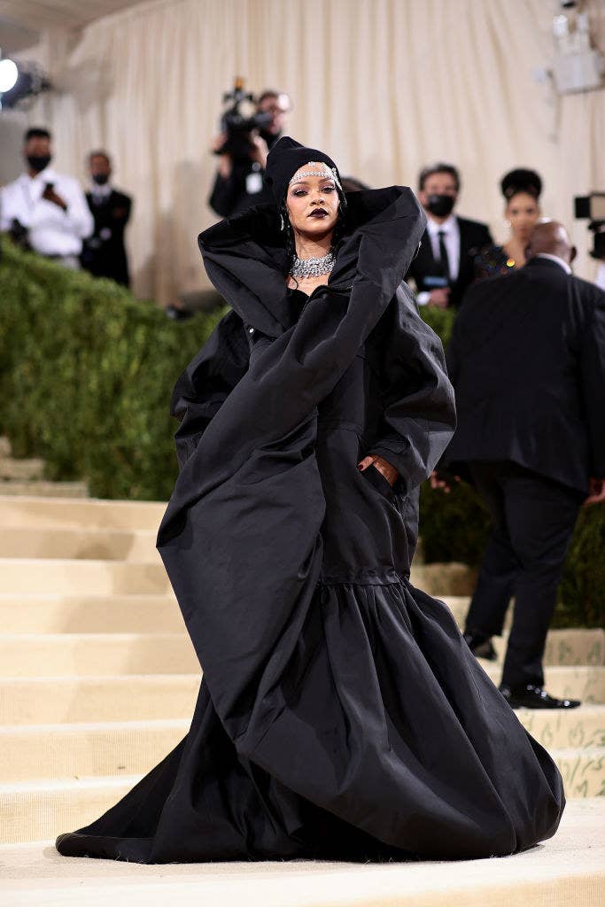 Rihanna wears a floor length dark gown with a high collar and a large diamond necklace, as well as a beanie