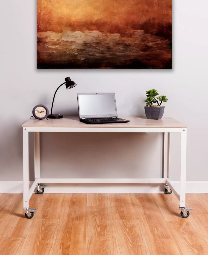 Wooden desk with white legs and wheels, lap top, lamp, and plant on desk top
