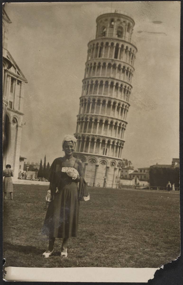 A woman wears a dress and floral headpiece in front of the Leaning Tower of Pisa in a midcentury photo