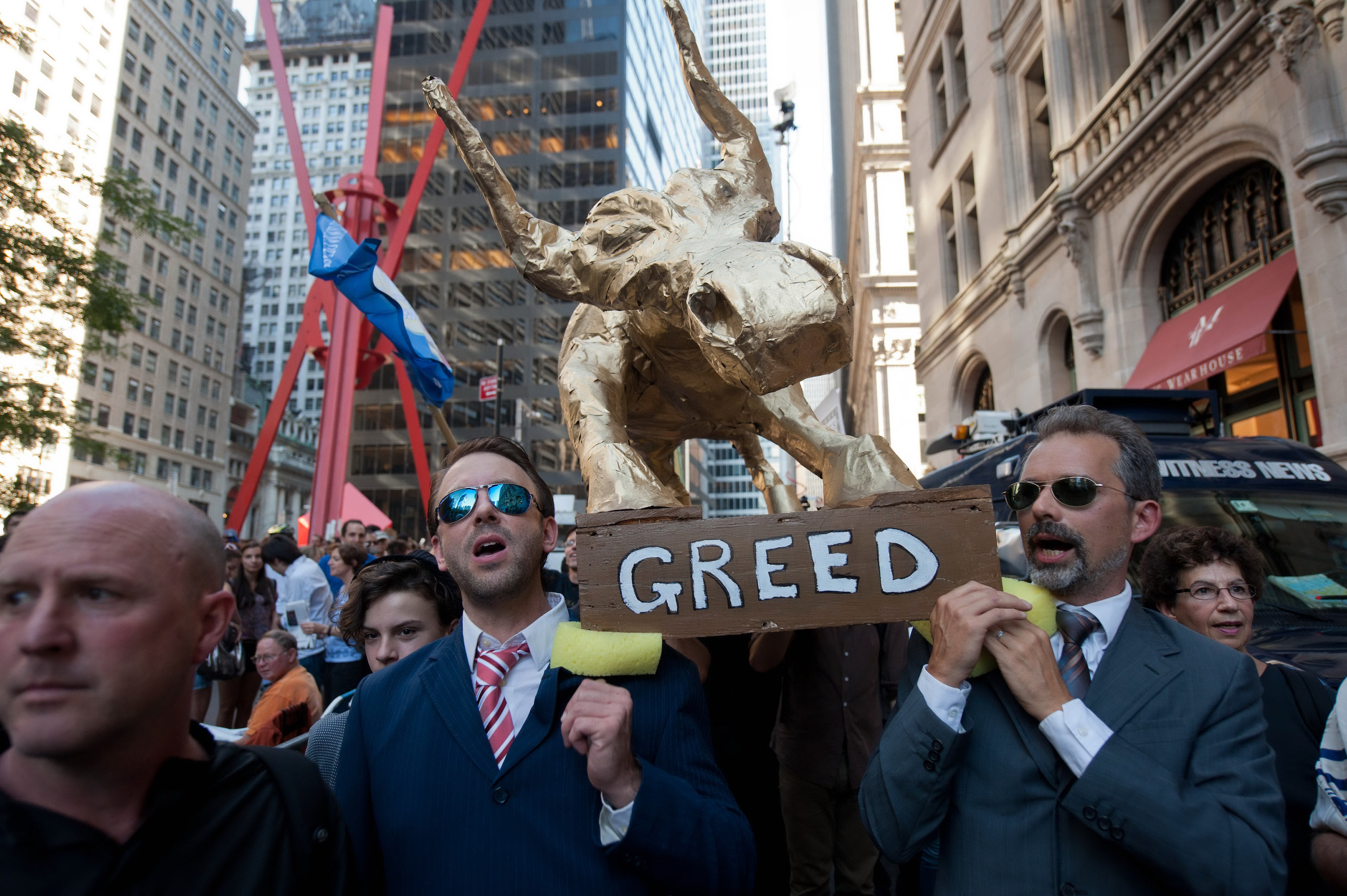 Two men in suits hold a sign that reads &#x27;greed&#x27; in downtown New York