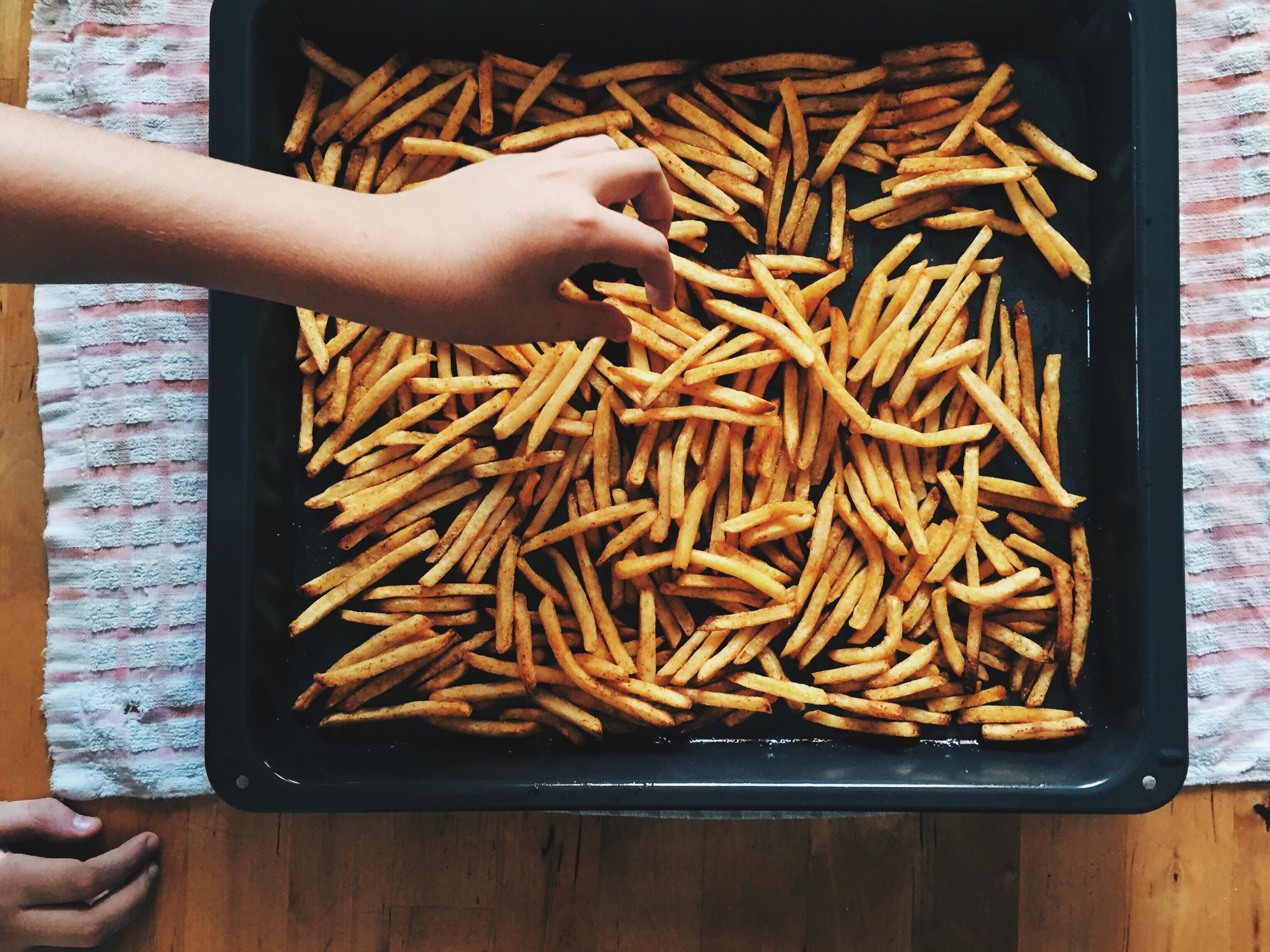 An overhead shot of fries in a baking tray and a hand taking out a fry.