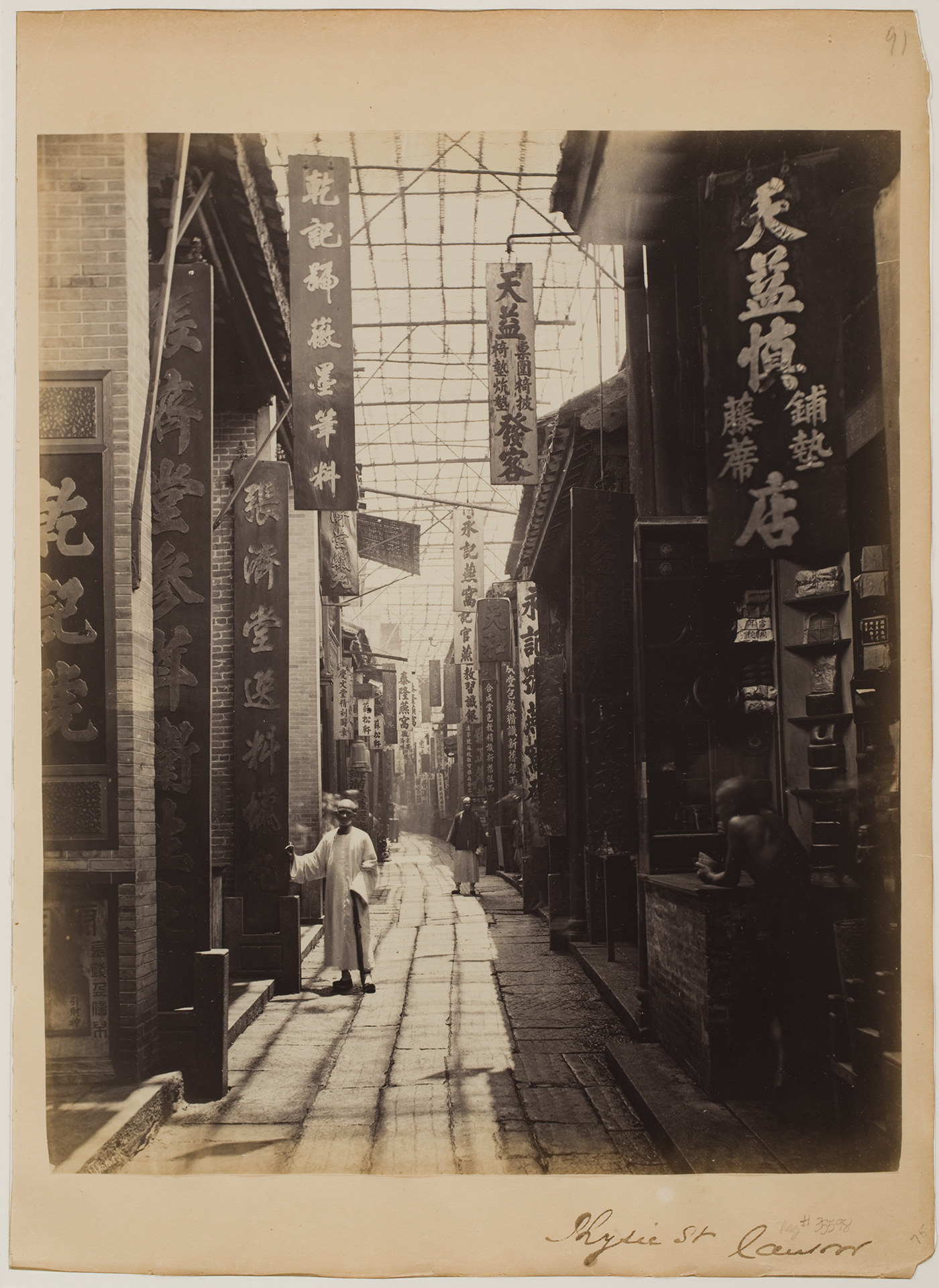 A man stands near a doorway in China near two other men, one standing in shadows, on an otherwise empty street of storefronts