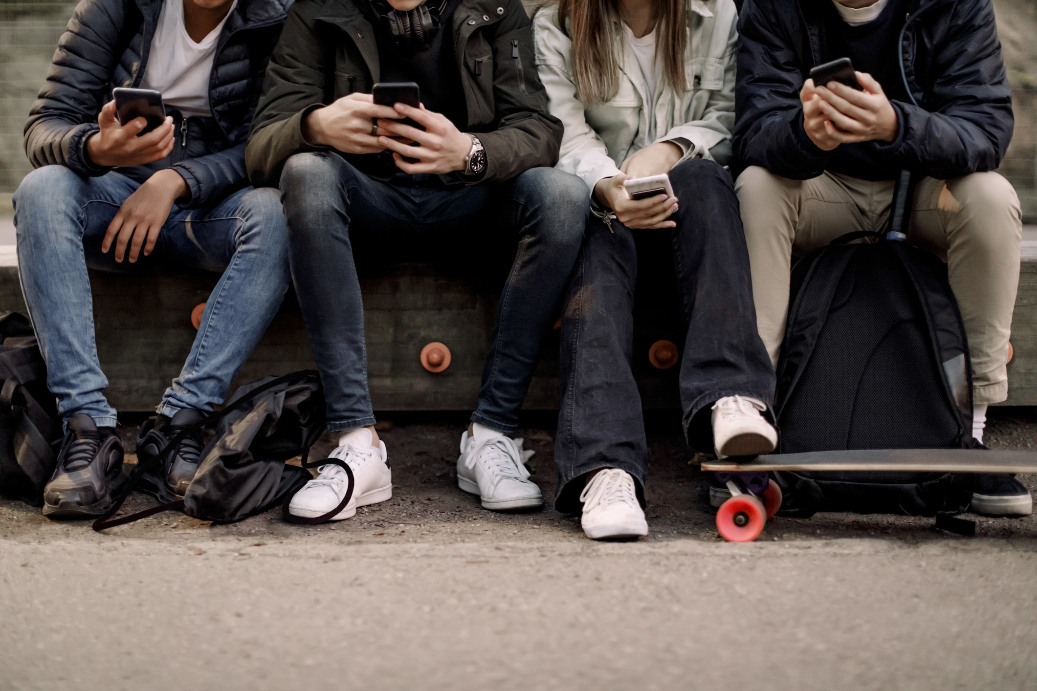 A group of teens, seen from the shoulders down, sitting together and hanging out outside looking at their phones
