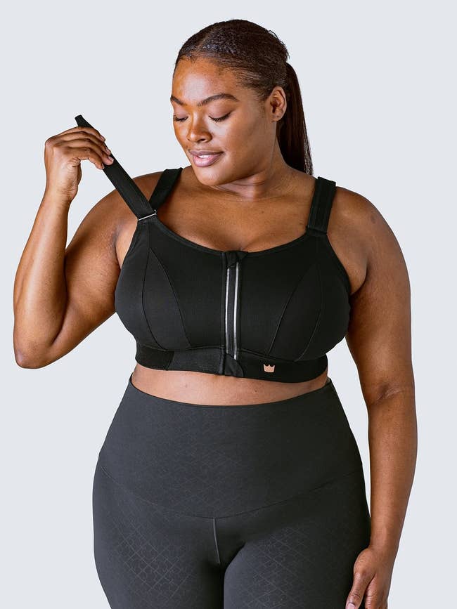 Model adjusting the sports bra in black with thick straps, a wide band around the chest and a zipper down the front