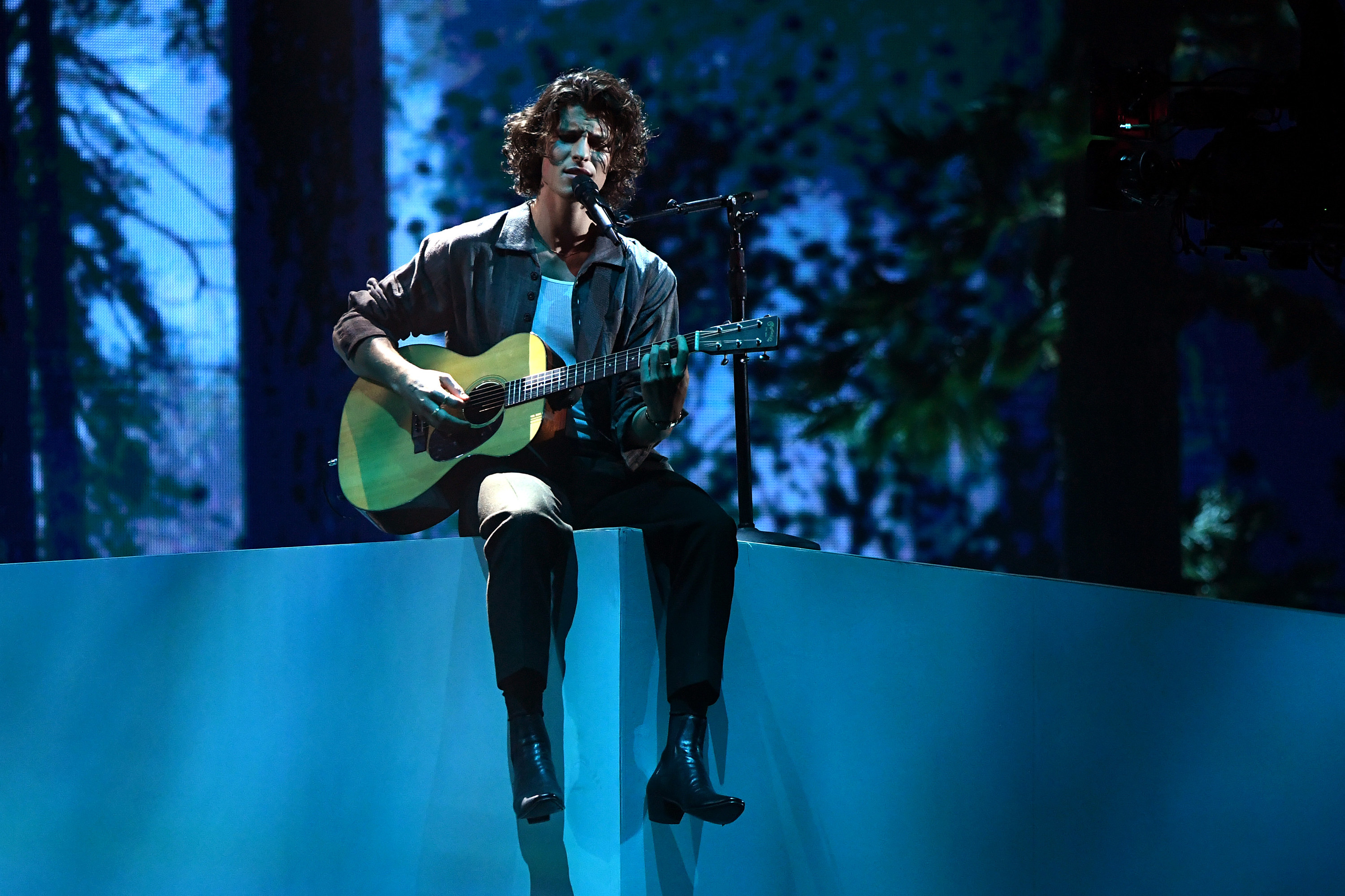 Shawn sits on a ledge on a stage while performing and playing guitar