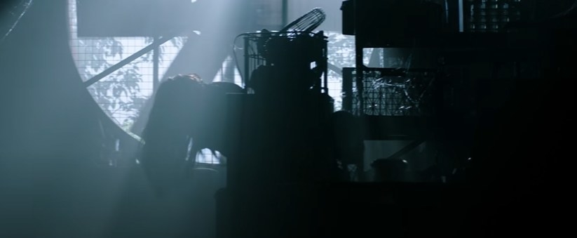 Long-haired figure leans over in front of a giant mechanical fan