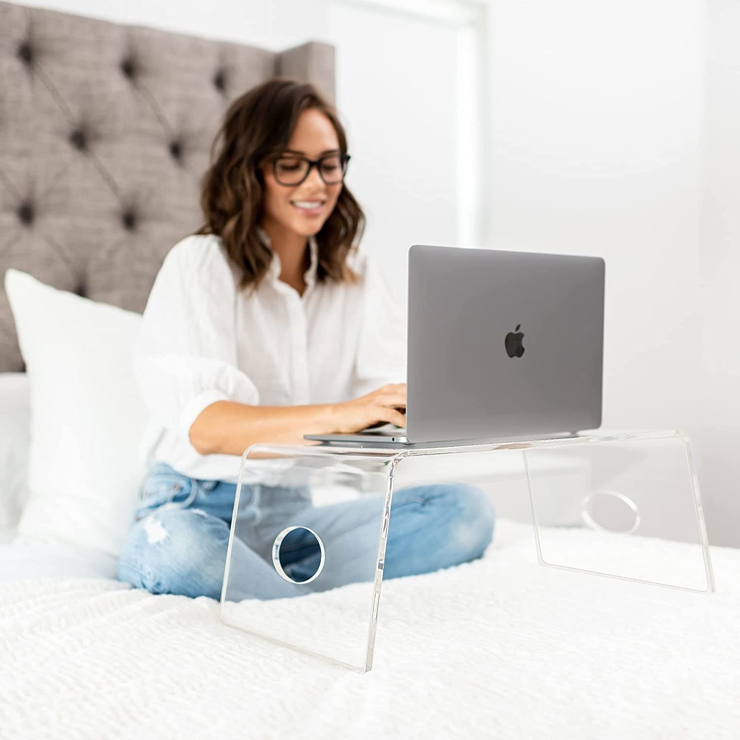 Model is sitting on a bed with a clear acrylic laptop table and a laptop on top of it
