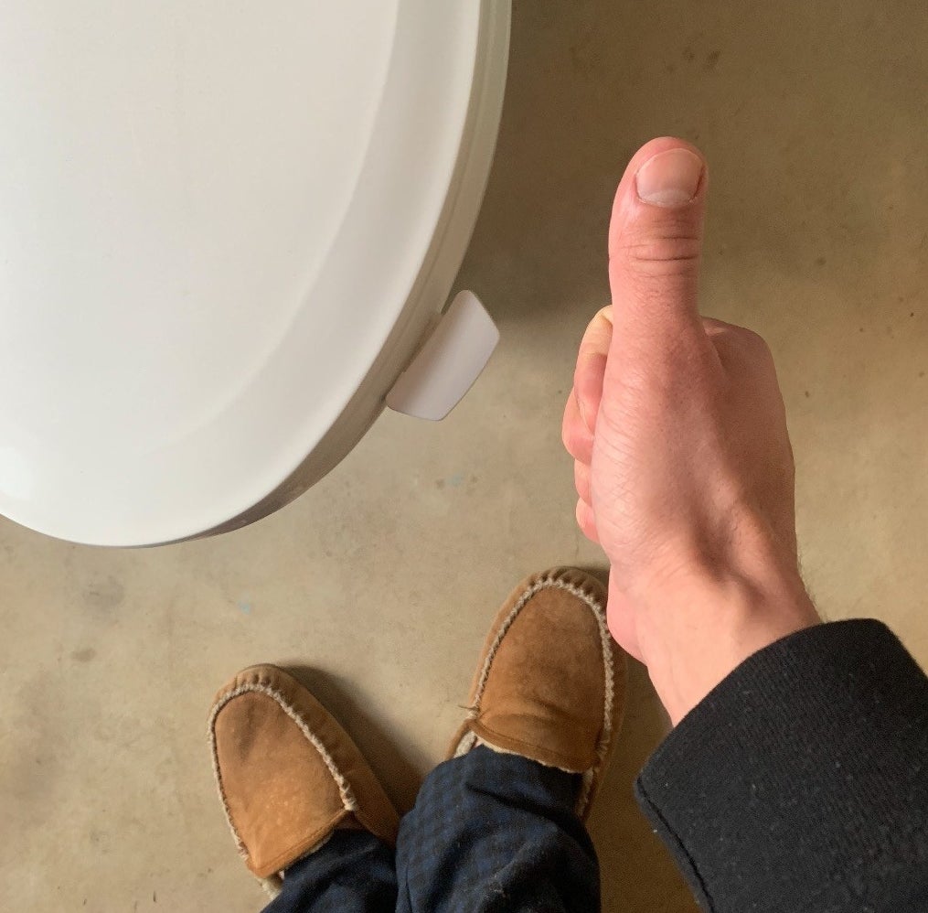 Reviewer is standing above a toilet with a lid lifter attached while giving a thumbs up