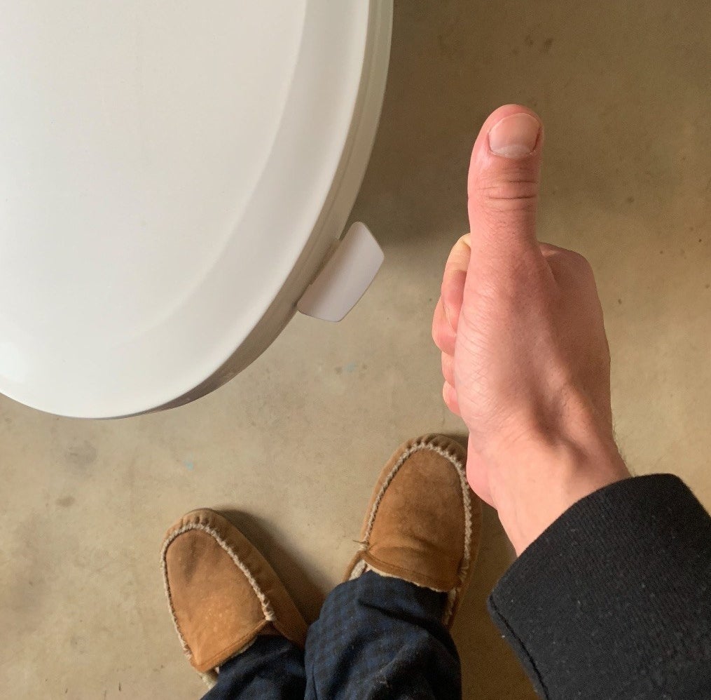 Reviewer is standing above a toilet with a lid lifter attached while giving a thumbs up