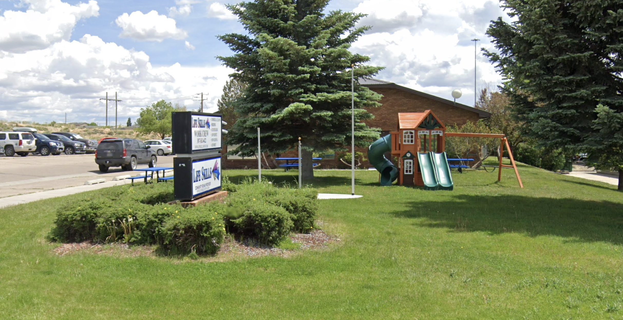 A grass lawn with a small playground and the &quot;Life Skills&quot; sign, near a road and parking lot 