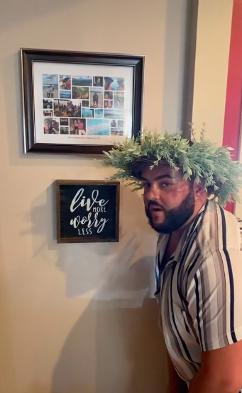 Man wearing a wreath and standing next to a &quot;Live more worry less&quot; sign on a wall below a framed montage of small photos