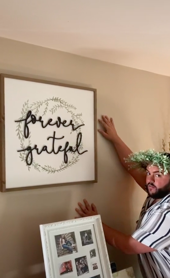Wreathed man touching a wall next to a framed &quot;Forever grateful&quot; wall hanging