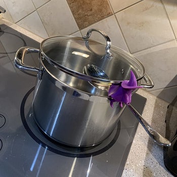 purple witch figure attached to side of reviewers pot, holding spoon like she's riding it as a broom and keeping the lid propped open