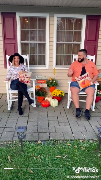 A woman wearing a large-brimmed hat and sitting on a porch with a &quot;Welcome autumn&quot; pillow in her lap, and a seated man smiling at her and holding a &quot;Blessed&quot; pumpkin pillow in his lap