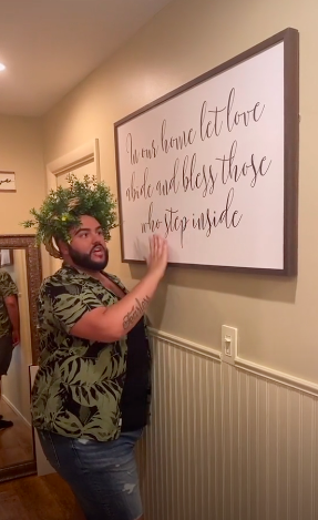 Wreathed man standing in front of a large framed wall hanging with the saying &quot;In our home let love abide and bless those who step inside&quot;