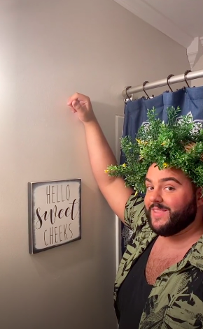 Smiling wreathed man showing off a framed &quot;Hello sweet cheeks&quot;  sign hanging on a wall