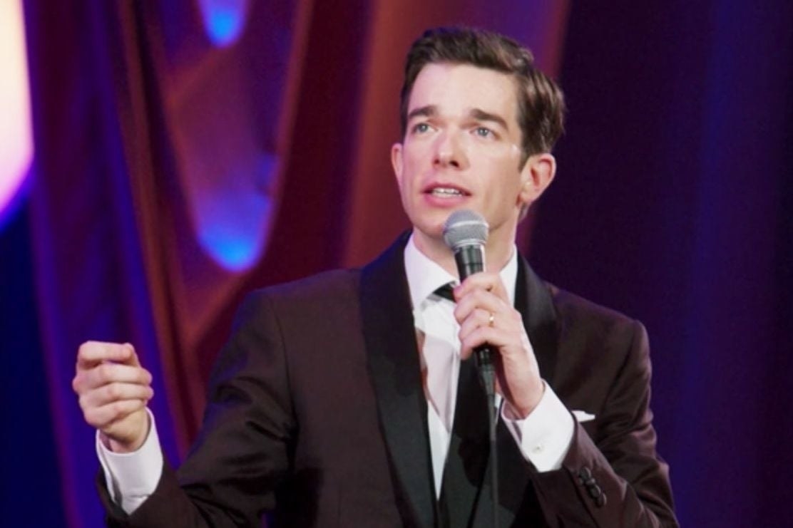 John Mulaney performing stand-up in a dark maroon suit