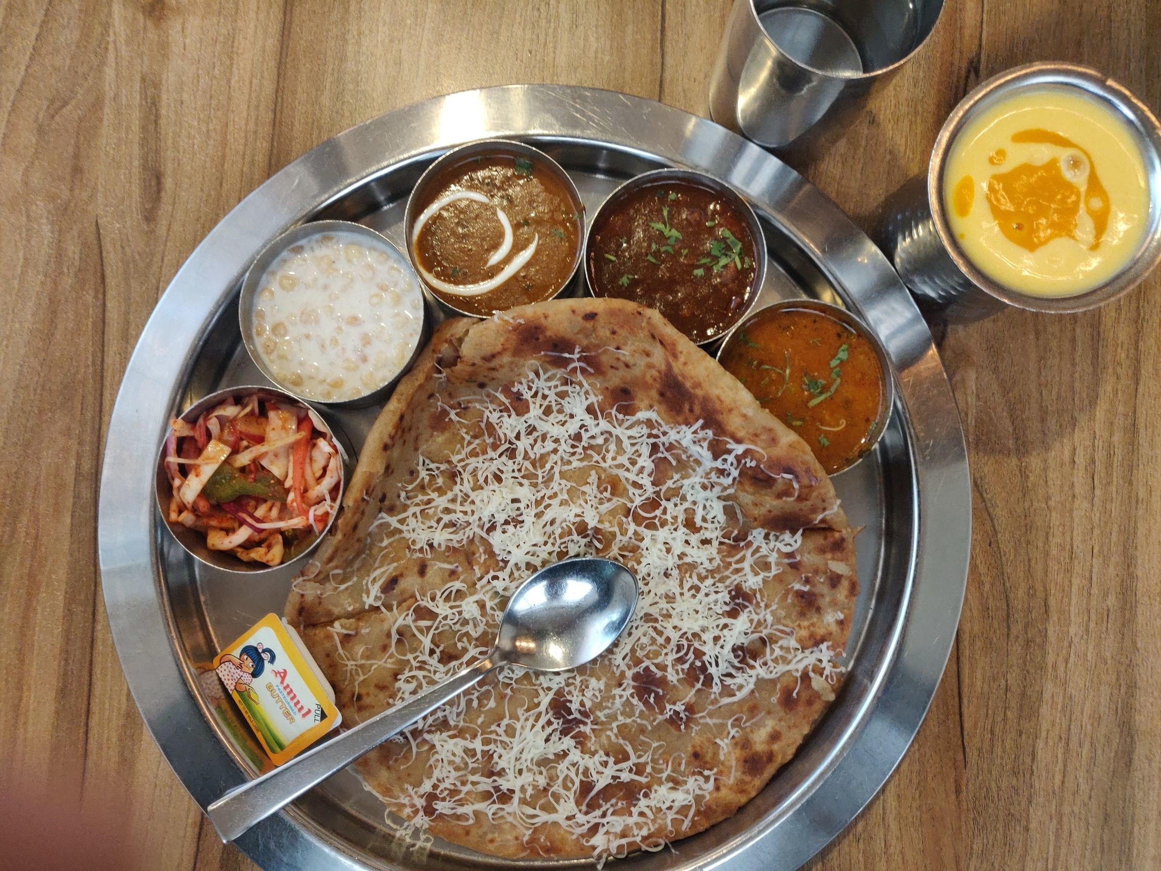 An aloo cheese paratha with dal makhni, chana masala, aloo sabzi, raita, salad, and butter. Glasses of water and mango lassi are kept on the side.