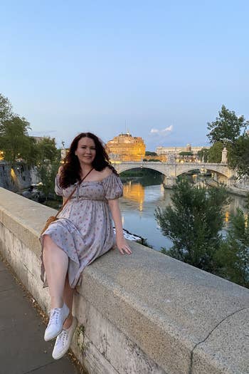 writer in a midi length dress with off the shoulder puff sleeves sitting on the wall of a bridge