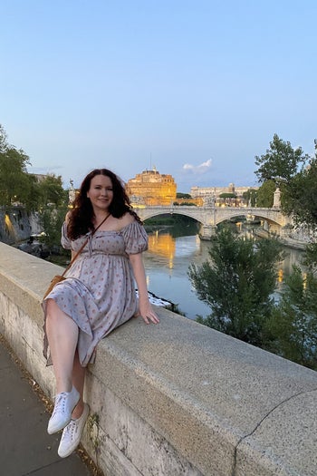 writer in a midi length dress with off the shoulder puff sleeves sitting on the wall of a bridge