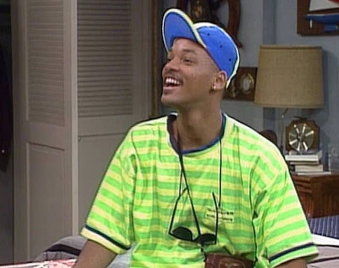 Will Smith wears a bright-color striped T-shirt and hat and sits on a bed smiling