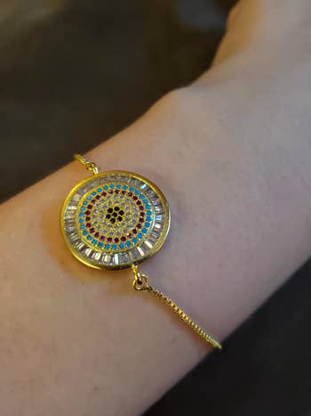 reviewer's wrist wearing the adjustable chain bracelet which has a gold charm in the center with colorful crystals on it arranged in a circle