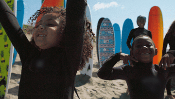 two kids in wet suits cheering on the beach with surf boards behind them