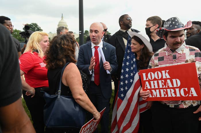 A number of people stand together holding signs and one person holding a flag, with the Capitol in the background