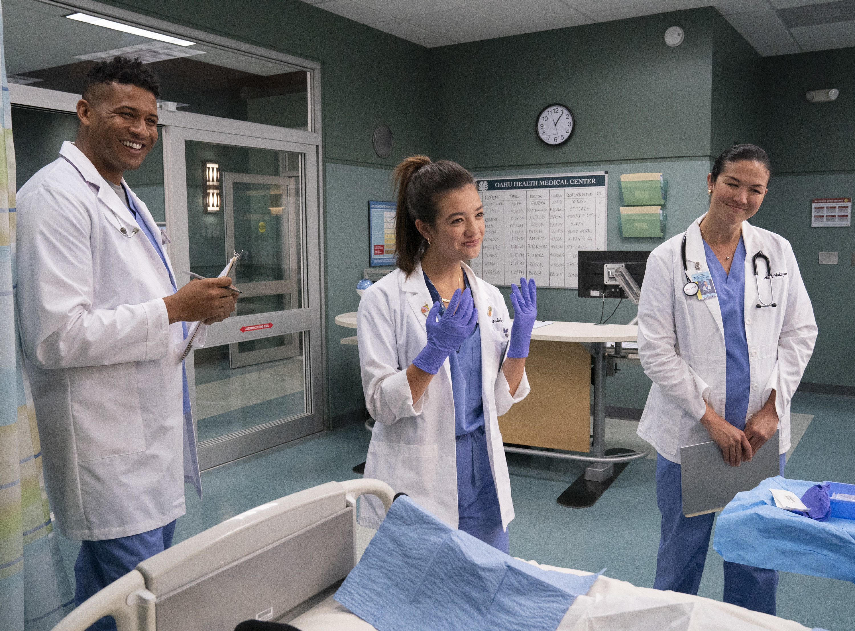 Jeffrey Bowyer-Chapman,  Peyton Elizabeth Lee, and Mapuana Makia stand near a hospital bed in doctor&#x27;s uniforms
