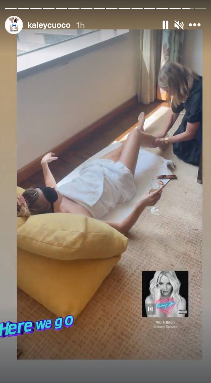 Kaley Cuoco lays on a mat with an eye covering while wearing a towel, holding a glass of wine, and getting her legs rubbed