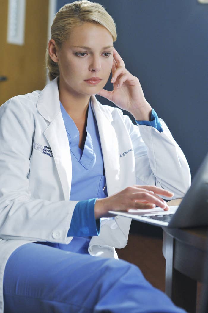 Heigl looks at a laptop with her hand touching her face while wearing scrubs