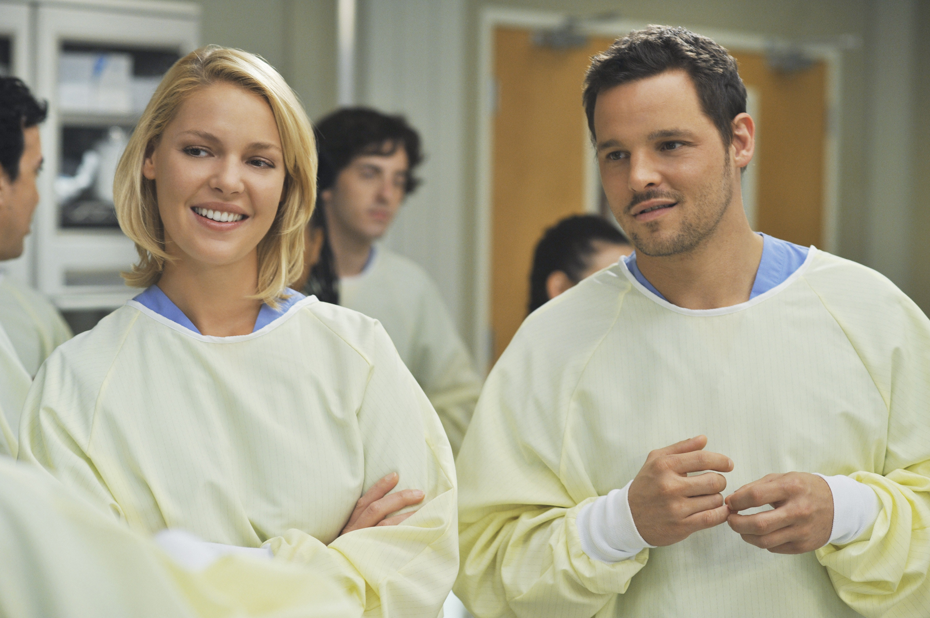 Heigl stands with her arms crossed next to another actor while wearing OR scrubs