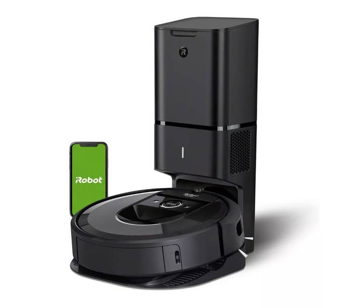 A black, iRobot Roomba vacuum with an automatic dirt disposal placed next to an iPhone with the iRobot app displayed
