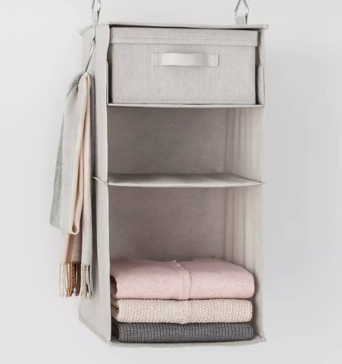 A light gray canvas, 3-tier hanging shelf filled with sweaters and scarfs hanging from the attached hook on the side