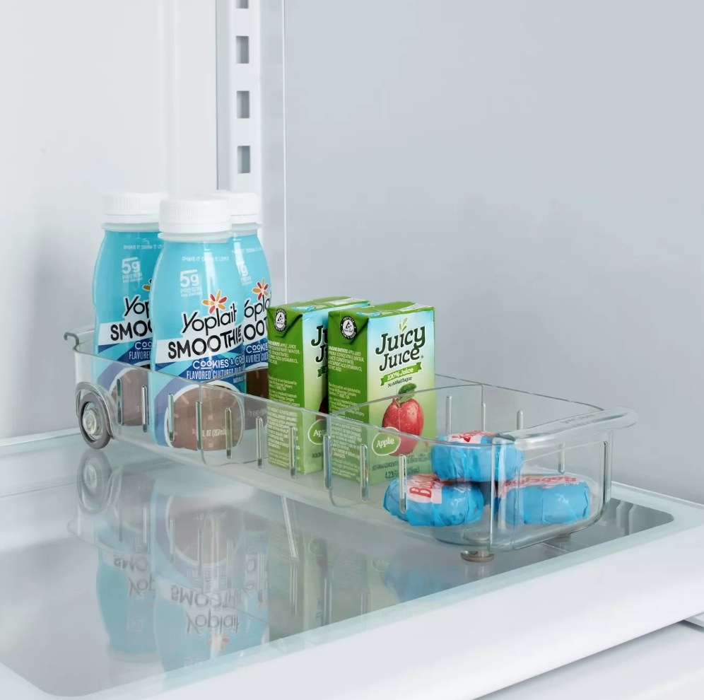 A clear fridge caddy with wheels in the back filled with juice boxed and cheese
