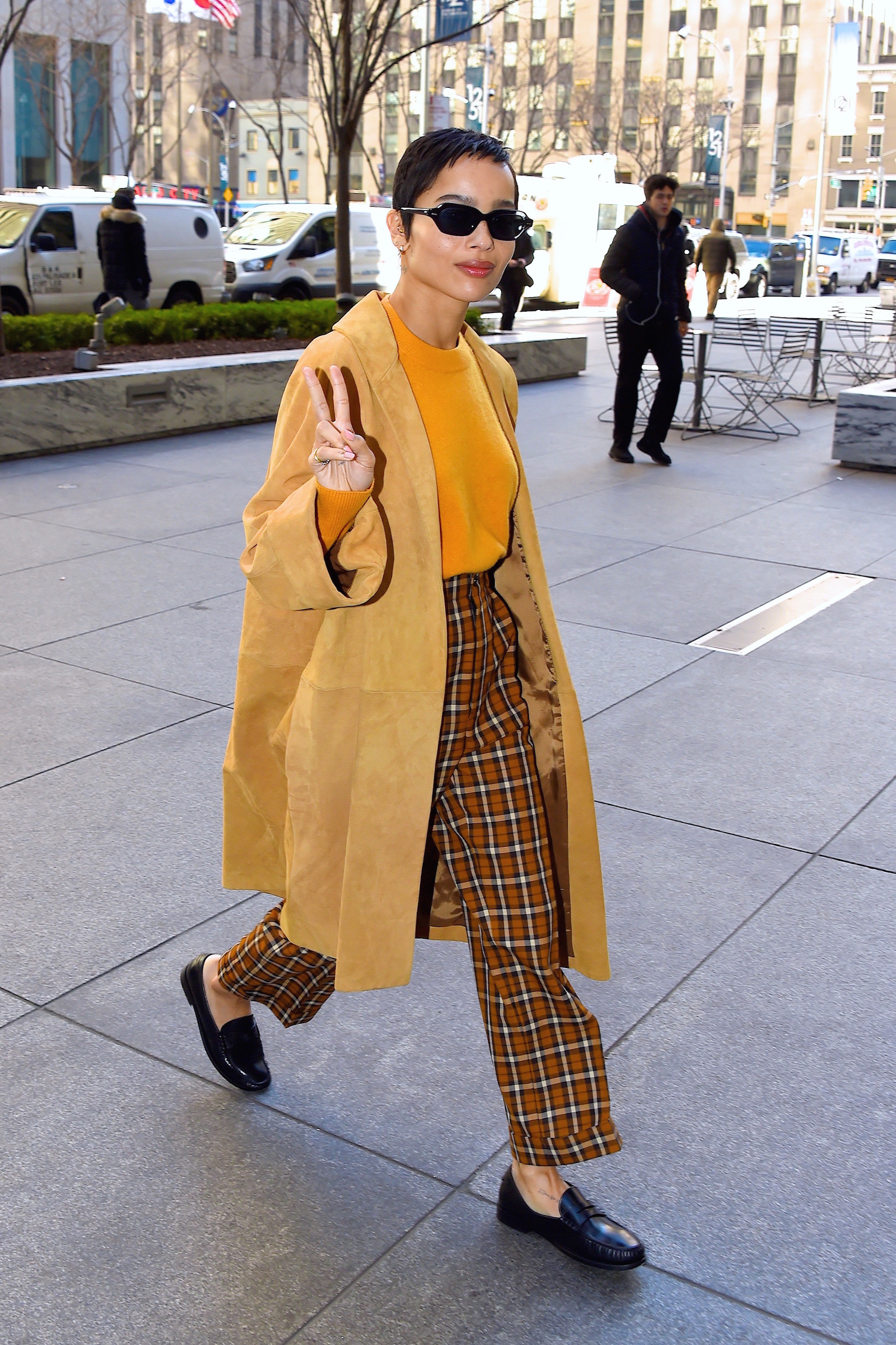 Zoe on a sidewalk in long jacket plaid pants loafers giving peace sign