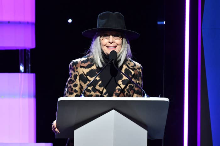 Diane speaking at a podium in an animal-print jacket, turtleneck, and fedora