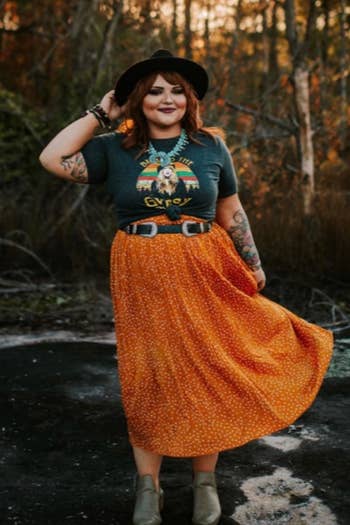 A reviewer wearing the mustard skirt with white dots paired with a belt, graphic tee, booties, a hat, and a statement necklace