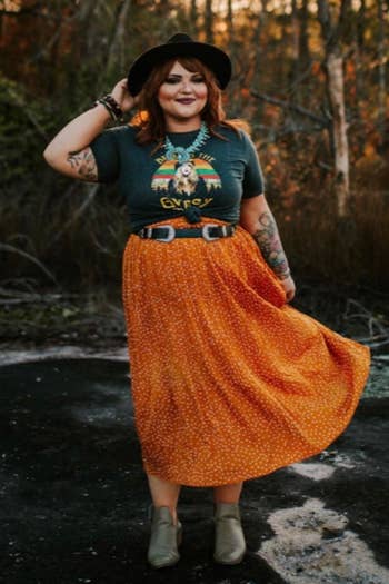 A reviewer wearing the mustard skirt with white dots paired with a belt, graphic tee, booties, a hat, and a statement necklace