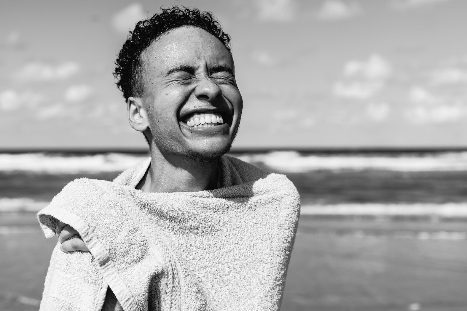 A young person smiles wide at the camera while wrapped in a towel, the ocean in the background 