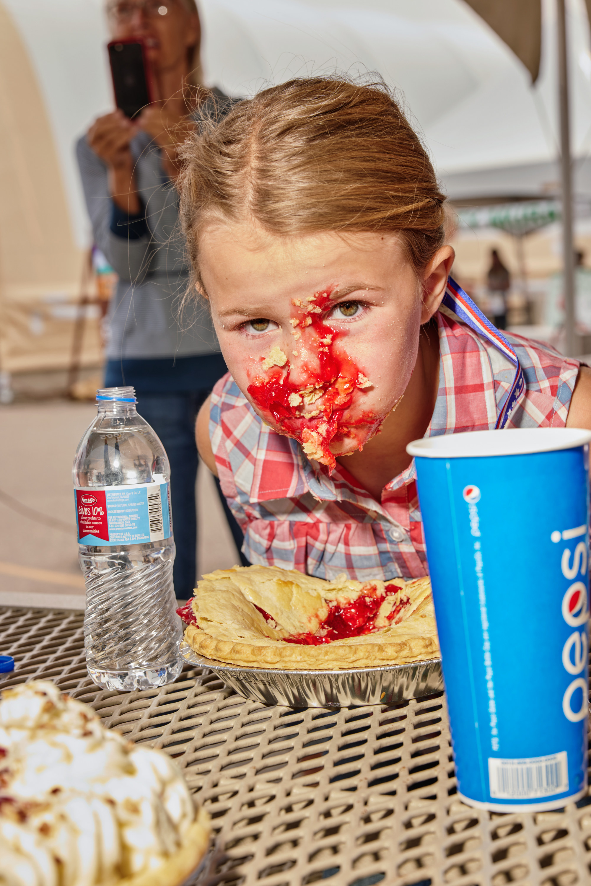 A girl lifts her face up from a pie, with pie filling all over her face, and a bottle of water to her left