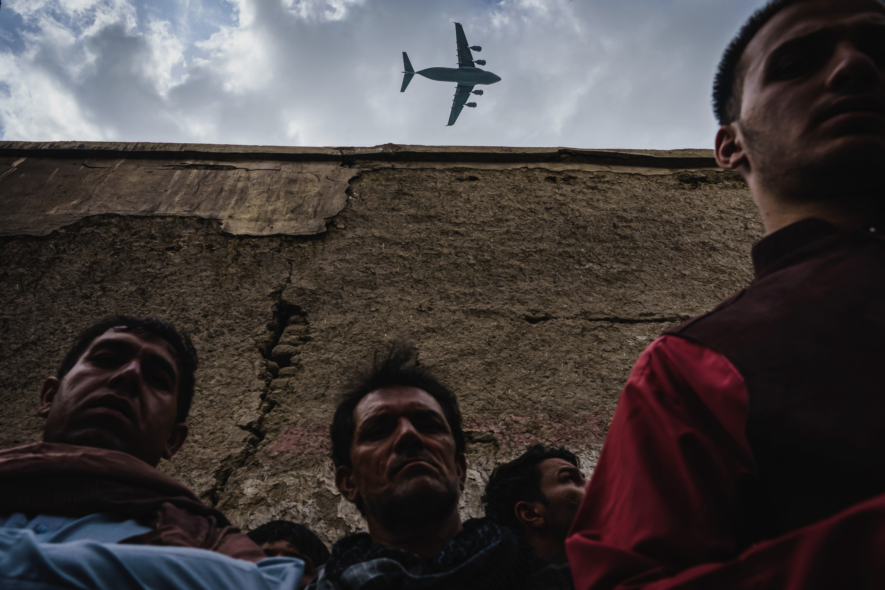 Three Afghan men stand against a wall, a plane flies overhead