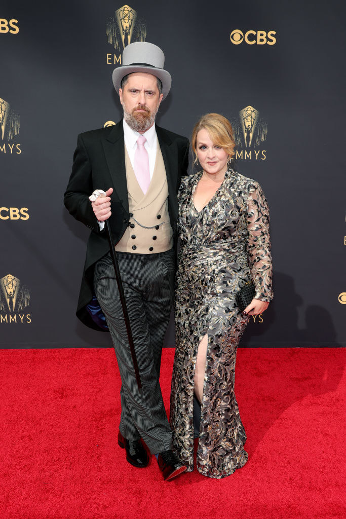 Brendan Hunt wears a dark suit, light colored vest, and a top hat, and Shannon Nelson wears a floral long sleeve gown with a slit up her thigh