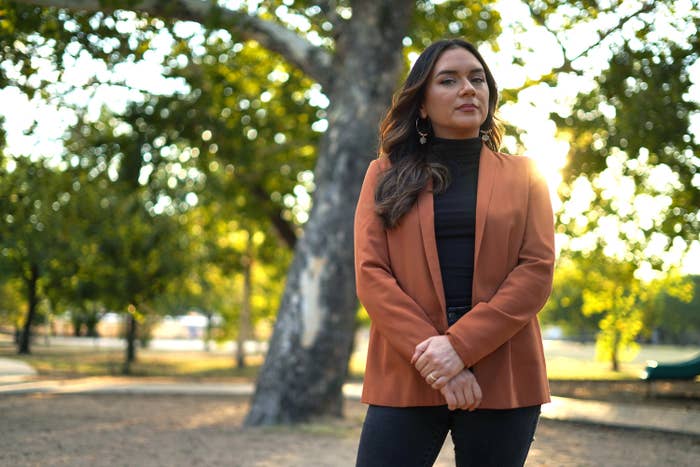 A woman in a blazer stands in front of a tree in the park, arms crossed and looking at the camera