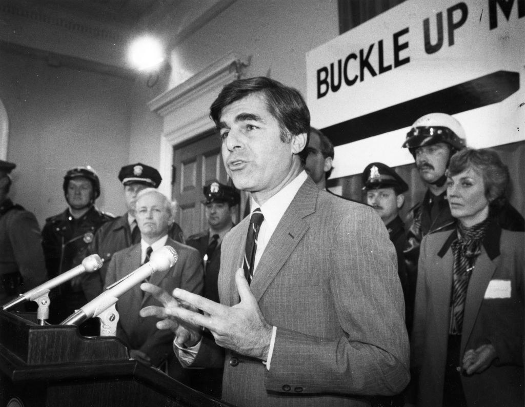 A man speaking at a podium with law enforcement behind him with a sign that says Buckle Up