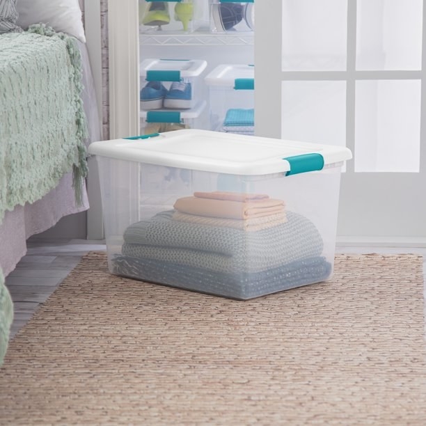 Storage bin with blankets inside, sitting on a rug next to a bed.