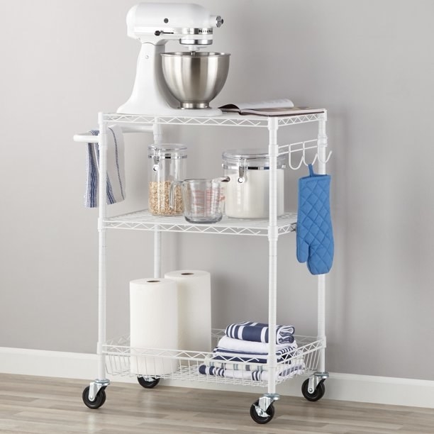 White wire cart holding a mixer, dish towels, paper towels, a measuring cup and glass canisters.